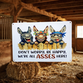 White aluminum sign with three grinning cartoon donkeys leaning over a wooden fence, reading “Don’t worry, be happy, we’re all asses here!”