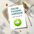 A white greeting card with bold dark teal text reading 'YOU'RE FUCKING AWESOME' and smaller text 'KEEP THAT SHIT UP,' plus a large green thumbs-up icon. The card is styled on cream fabric with dried grass and small shells in soft natural light.