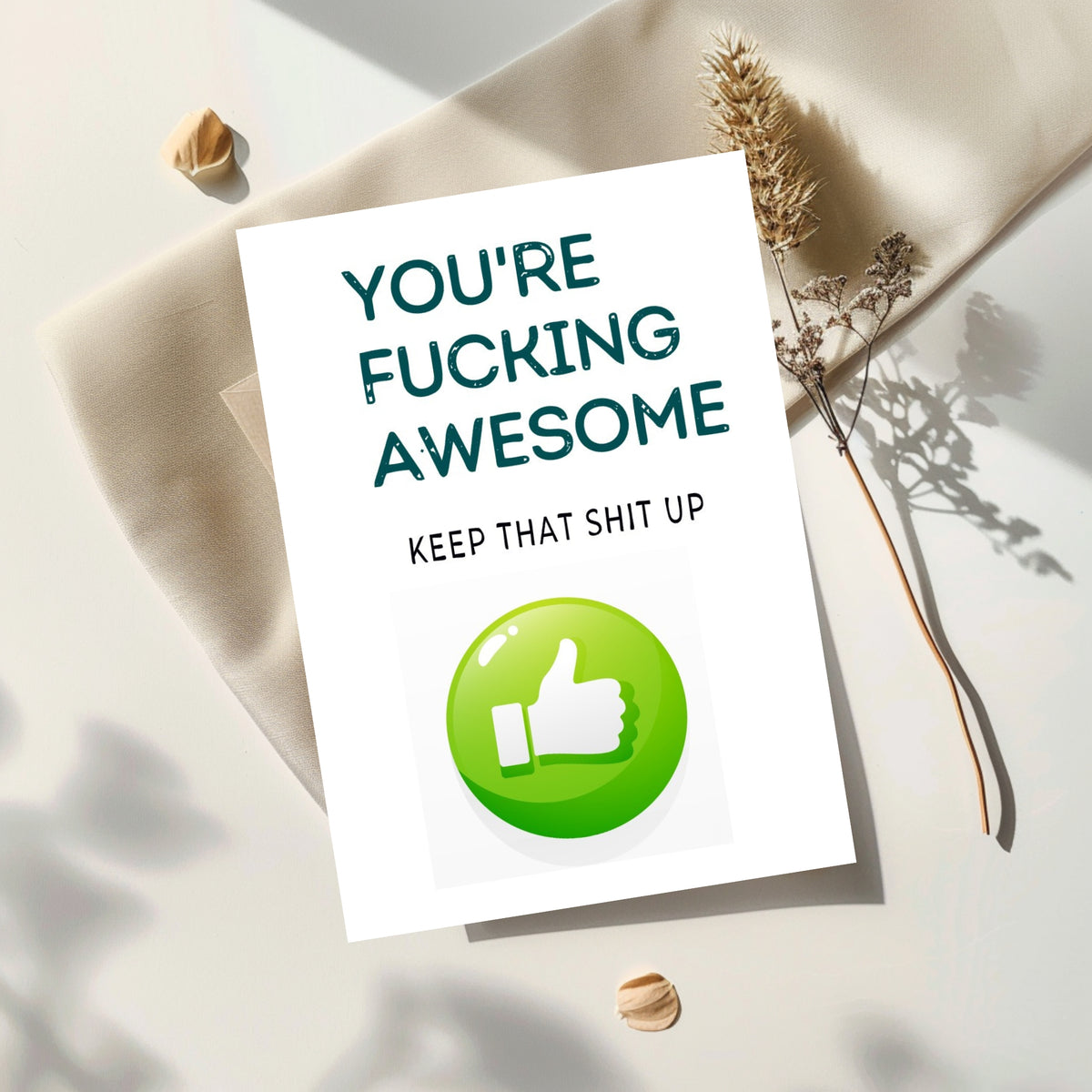 A white greeting card with bold dark teal text reading 'YOU'RE FUCKING AWESOME' and smaller text 'KEEP THAT SHIT UP,' plus a large green thumbs-up icon. The card is styled on cream fabric with dried grass and small shells in soft natural light.
