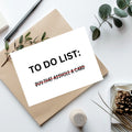 A white to-do list card with black and red text reading 'TO DO LIST: BUY THAT ASSHOLE A CARD' is placed on top of a kraft paper envelope, styled with eucalyptus leaves, a pen, and a small decorative object on a white surface.