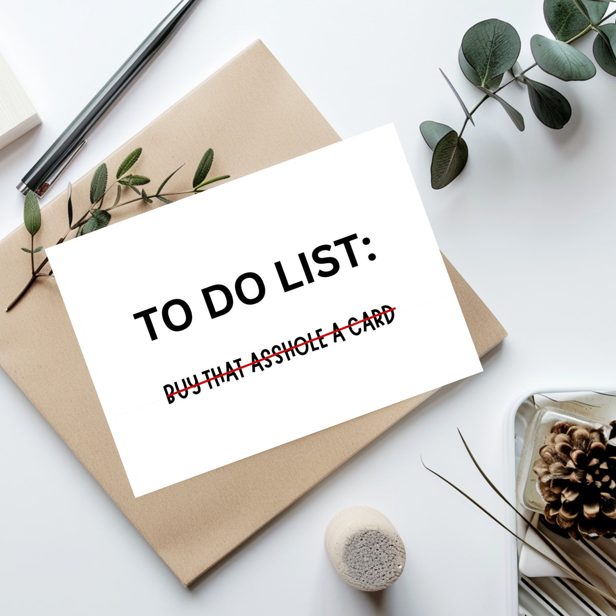 A white to-do list card with black and red text reading 'TO DO LIST: BUY THAT ASSHOLE A CARD' is placed on top of a kraft paper envelope, styled with eucalyptus leaves, a pen, and a small decorative object on a white surface.