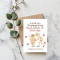 White rectangular card with a nude cartoon older couple and red hearts, reading “Love is, supporting each other in old age” and “Happy Anniversary.”