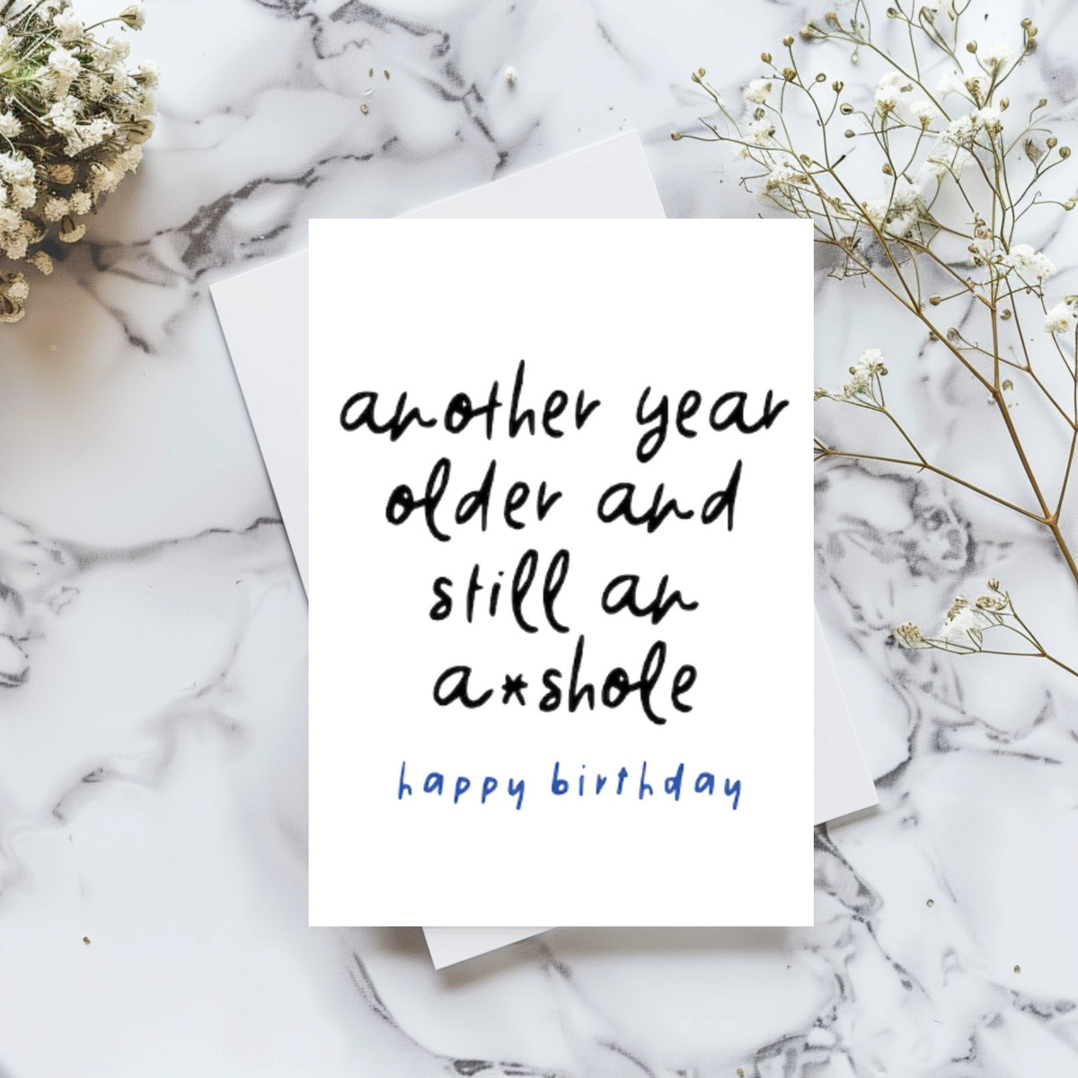 A white birthday card with black handwritten text reading 'another year older and still an a*shole' and blue text reading 'happy birthday' underneath, styled on a white marble surface with baby's breath flowers.