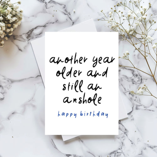 A white birthday card with black handwritten text reading 'another year older and still an a*shole' and blue text reading 'happy birthday' underneath, styled on a white marble surface with baby's breath flowers.