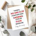 A white greeting card with bold red and black text reading 'THERE'S NO ONE ELSE I'D RATHER HAVE SNORING LOUD AS FUCK BESIDE ME. Happy Anniversary!' placed on a kraft envelope, styled with eucalyptus leaves and a wooden block on a white surface.