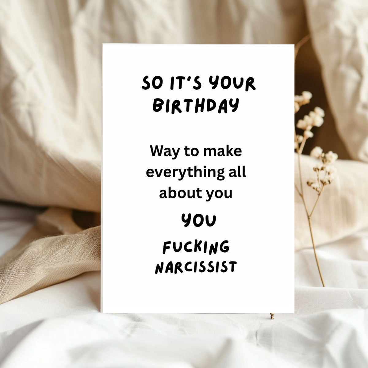 A white greeting card with black text reading 'SO IT'S YOUR BIRTHDAY Way to make everything all about you YOU FUCKING NARCISSIST' standing on white fabric with beige bedding and dried flowers in the background.