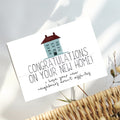 A white congratulations card featuring a small house illustration and the text 'CONGRATULATIONS ON YOUR NEW HOME! i hope your new neighbours aren't assholes', styled on a light surface with a wicker basket edge and dried stems nearby.