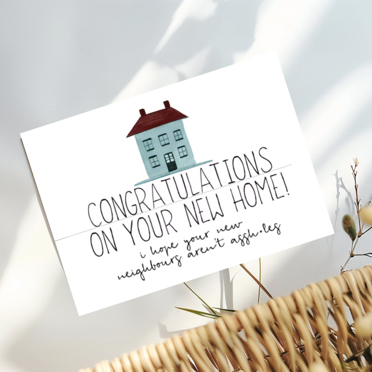 A white congratulations card featuring a small house illustration and the text 'CONGRATULATIONS ON YOUR NEW HOME! i hope your new neighbours aren't assholes', styled on a light surface with a wicker basket edge and dried stems nearby.