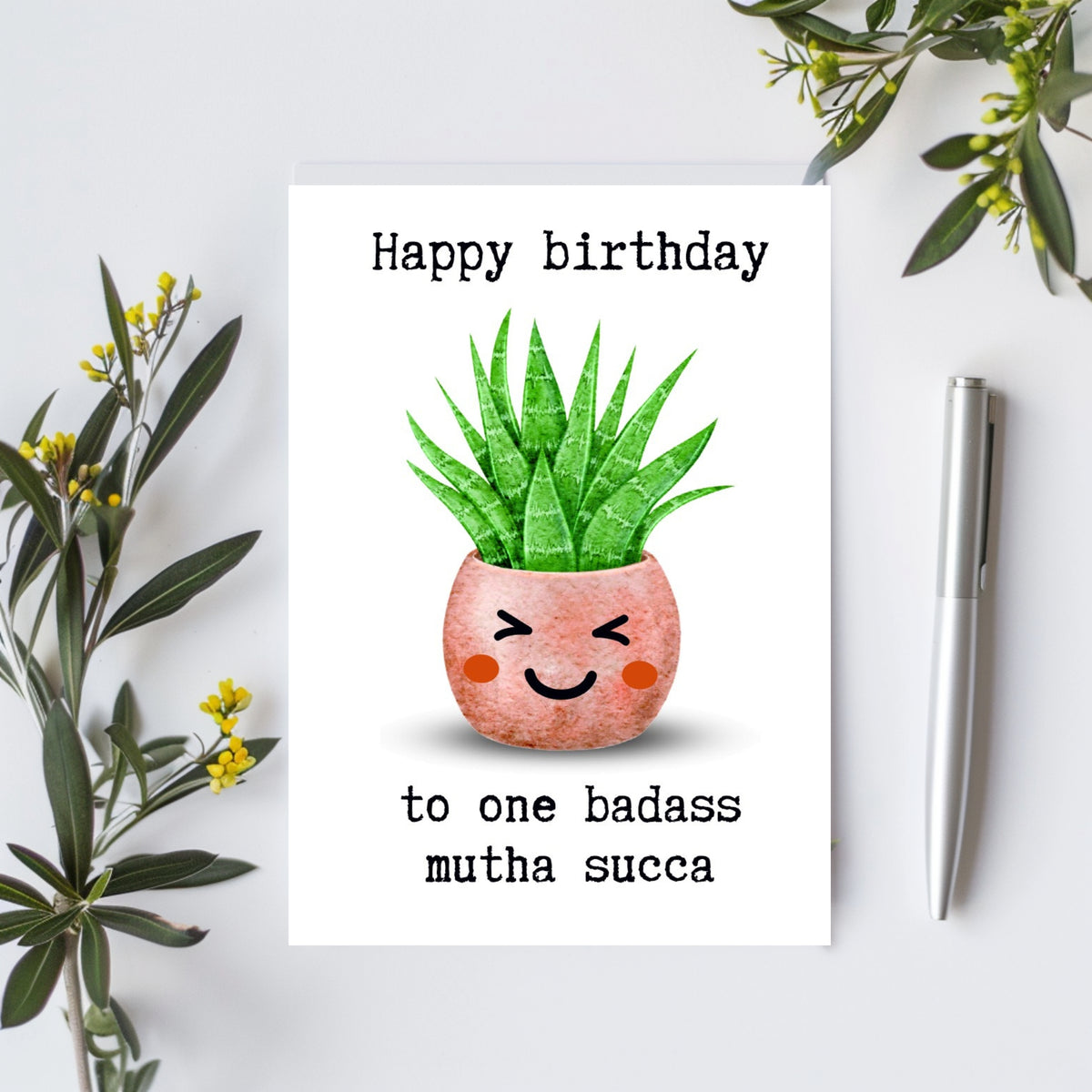 A greeting card featuring a cute cartoon succulent plant with a smiling face in a pink pot, with the text 'Happy birthday to one badass mutha succa'. The card is styled in a flat-lay setting on a white surface with green leaves and a silver pen.