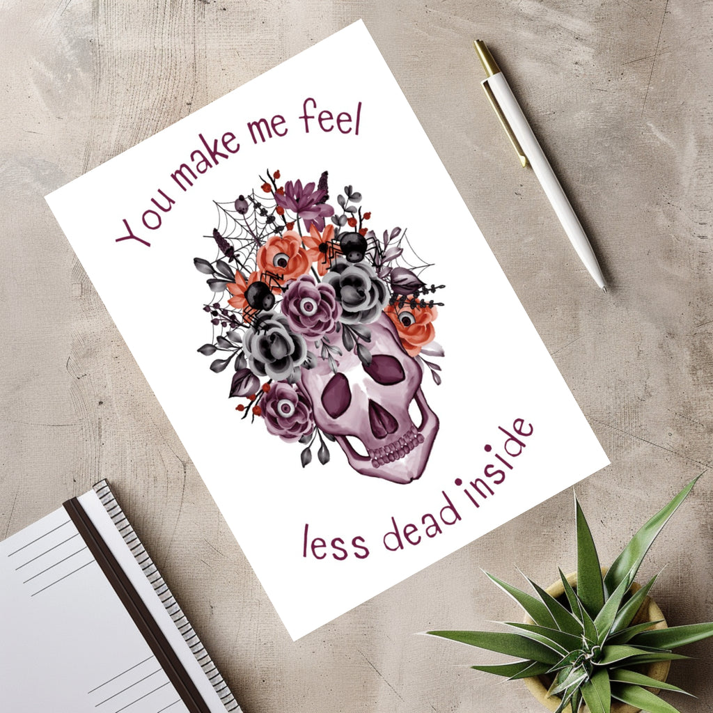 White rectangular greeting card with a purple skull decorated with flowers, spiders and webs, reading “You make me feel less dead inside.”