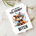 A white birthday greeting card featuring a cartoon grumpy cat tangled in Christmas lights, with the text 'HAPPY BIRTHDAY YOU GRUMPY BITCH'. The card is displayed on a marble surface with small white flowers as props.