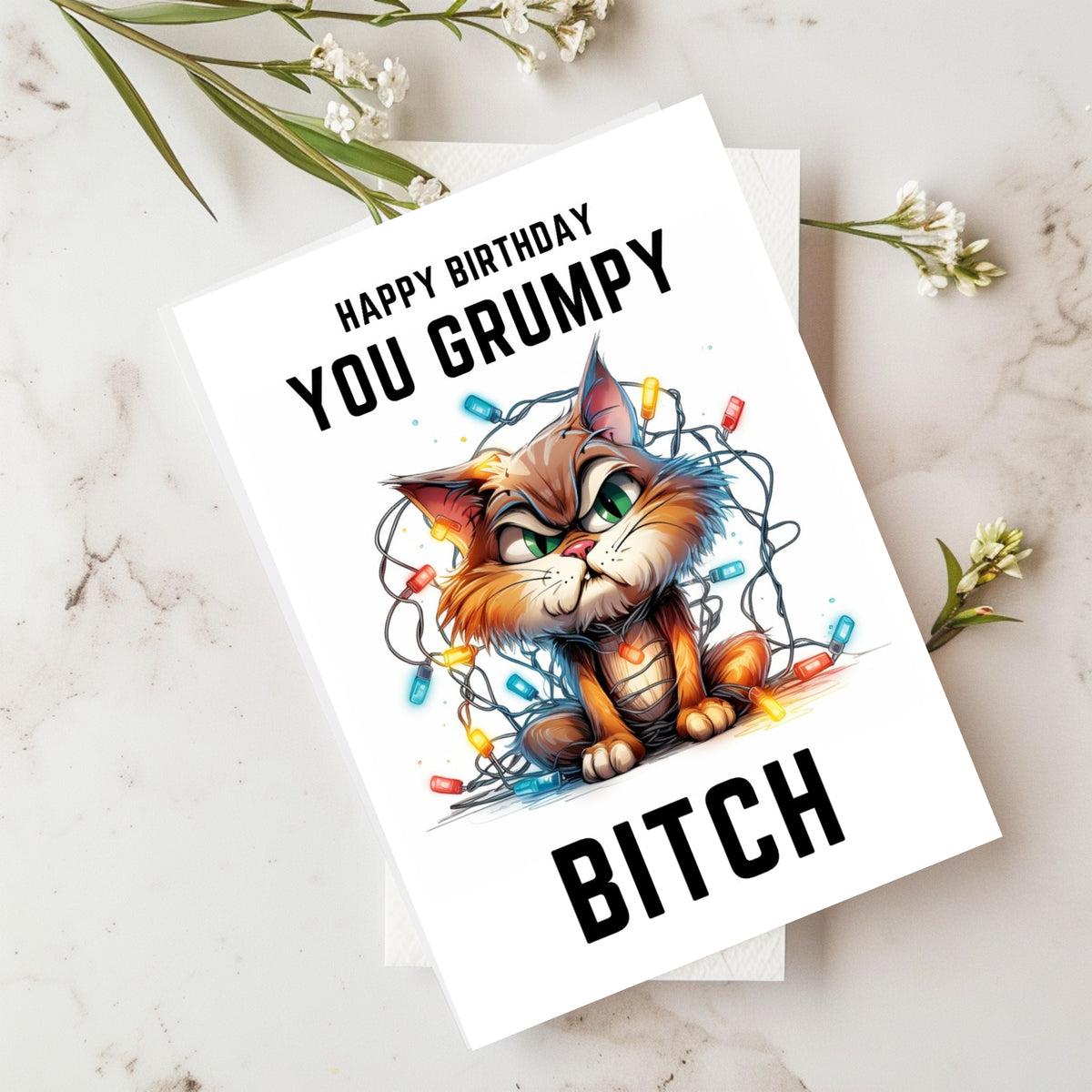 A white birthday greeting card featuring a cartoon grumpy cat tangled in Christmas lights, with the text 'HAPPY BIRTHDAY YOU GRUMPY BITCH'. The card is displayed on a marble surface with small white flowers as props.