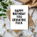 A white greeting card with bold black text reading 'HAPPY BIRTHDAY YOU GERIATRIC FUCK' is styled in a flat lay on burlap and twine over a whitewashed wood surface with green leaves and tan fabric around it.