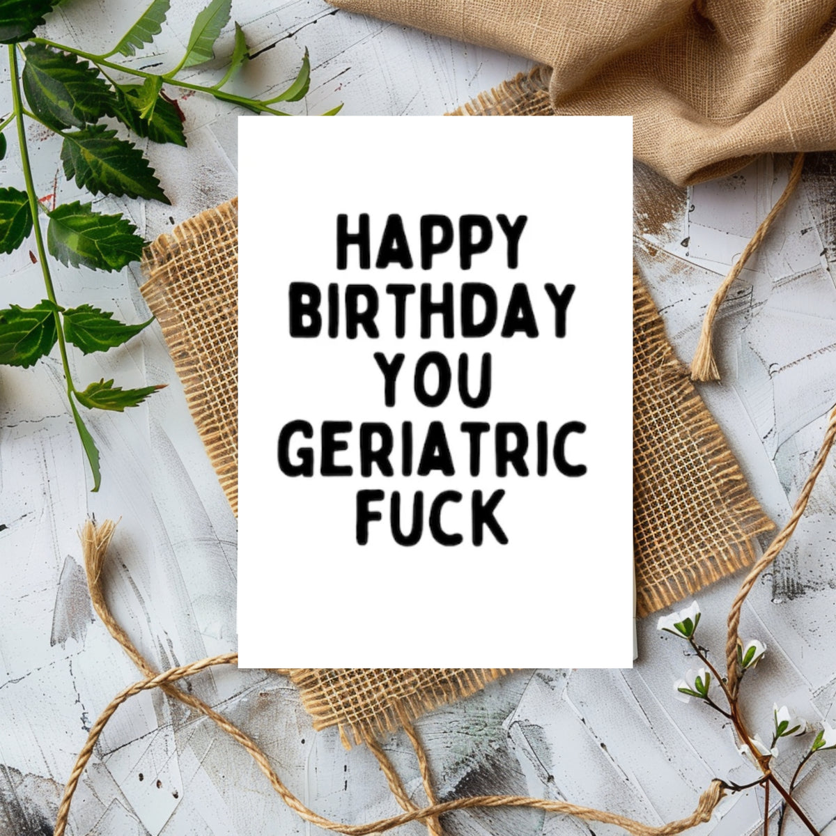 A white greeting card with bold black text reading 'HAPPY BIRTHDAY YOU GERIATRIC FUCK' is styled in a flat lay on burlap and twine over a whitewashed wood surface with green leaves and tan fabric around it.