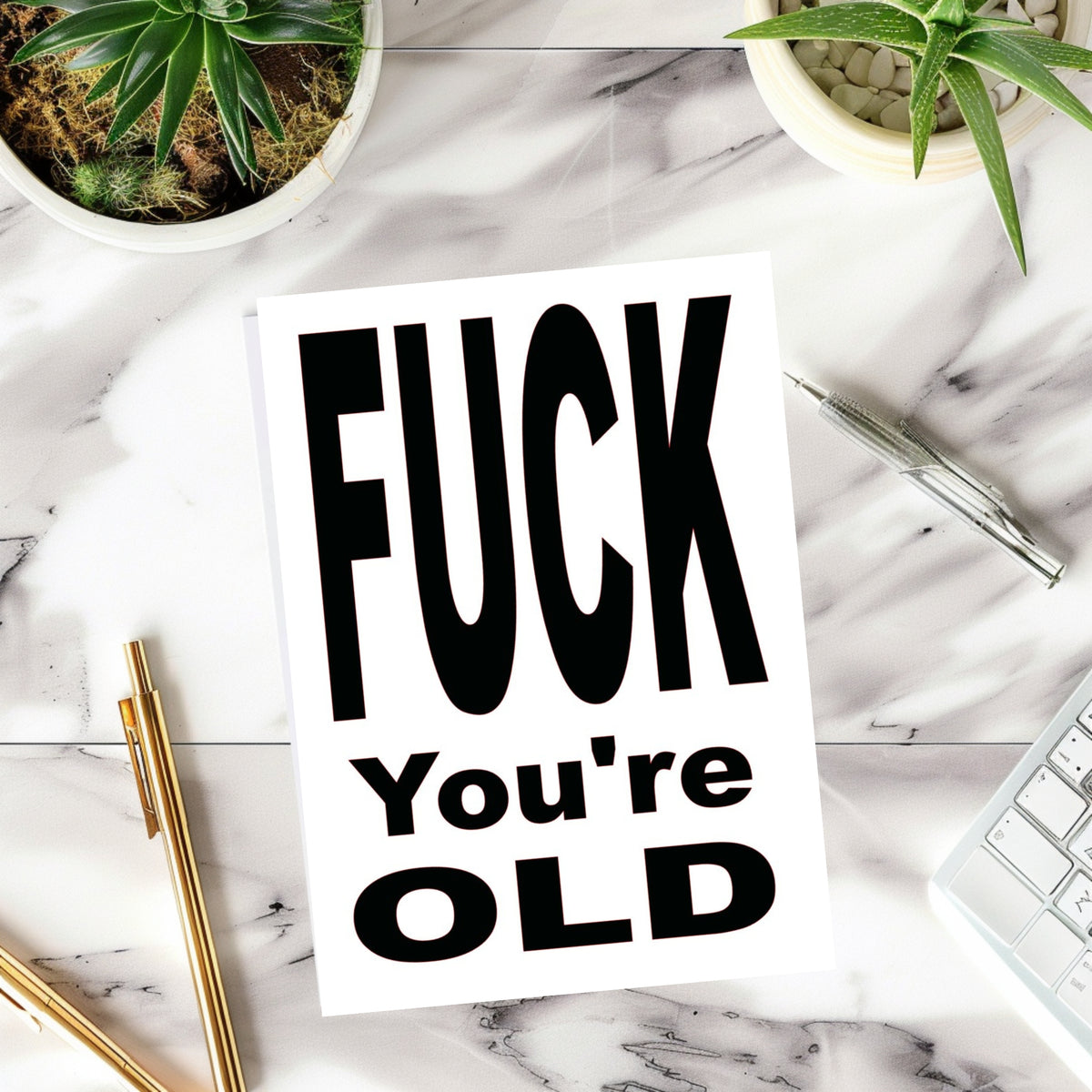 A white rectangular print or card with bold black text reading 'FUCK You're OLD' is placed on a white marble surface with small potted plants, pens, and part of a keyboard visible around it.