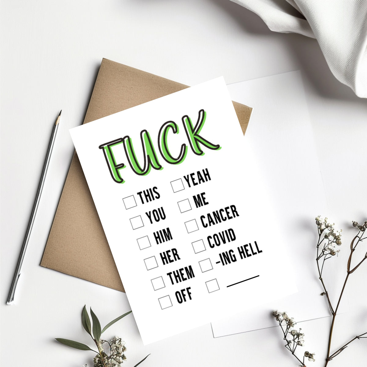 A white novelty greeting card with the word 'FUCK' in large green outlined letters at the top, followed by a checklist of phrases including 'THIS', 'YOU', 'HIM', 'HER', 'THEM', 'ME', 'YEAH', 'CANCER', 'COVID', 'ING HELL', and 'OFF'. The card is styled in a flat lay with a brown kraft envelope, a silver pen, white fabric, and small dried flowers on a white background.