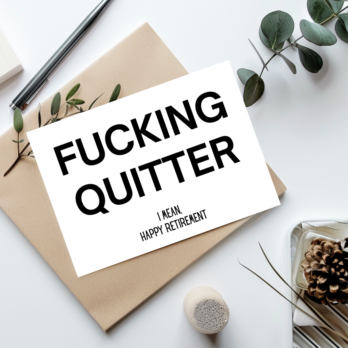 A white greeting card with bold black text reading 'FUCKING QUITTER' and smaller text 'I MEAN, HAPPY RETIREMENT,' styled on top of a kraft envelope with eucalyptus leaves, a pen, and other flat-lay props on a white surface.