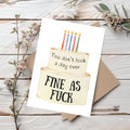 A greeting card featuring a two-tier birthday cake with colorful candles and the text 'You don't look a day over' and 'FINE AS FUCK', shown on a rustic white wood background with a kraft envelope and small dried flowers.