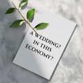 A white rectangular card or print with the text 'A WEDDING? IN THIS ECONOMY?' in black serif lettering, placed on a light gray textured surface with a green leafy stem across the top left.