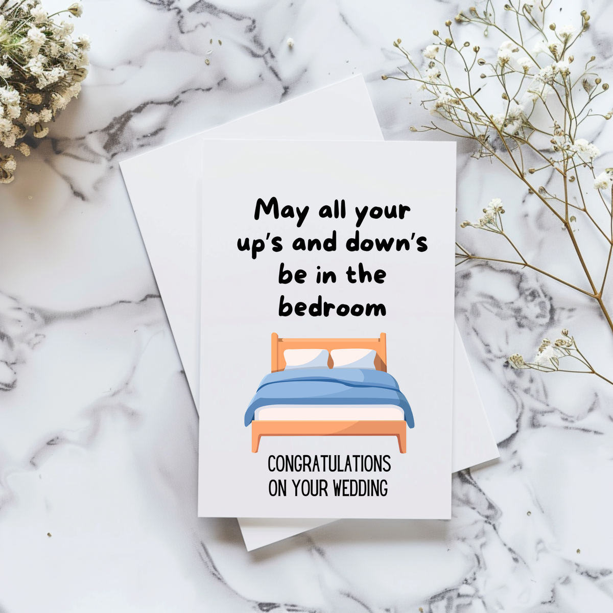 White rectangular wedding card with a wooden bed and blue bedding, reading “May all your up’s and down’s be in the bedroom” and “Congratulations on your wedding.”
