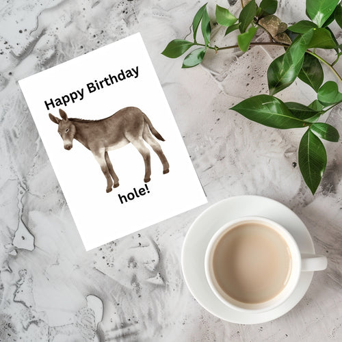 A white greeting card with a donkey illustration and the text 'Happy Birthday hole!' is styled on a light stone surface next to a white cup of coffee and green leaves.