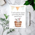 A baby shower greeting card featuring a watercolor elephant in a wooden basket with the text 'I can't wait to teach your baby its first swear word', styled in a flat lay on a white marble surface with green leaves, a pen, and a stone tile.