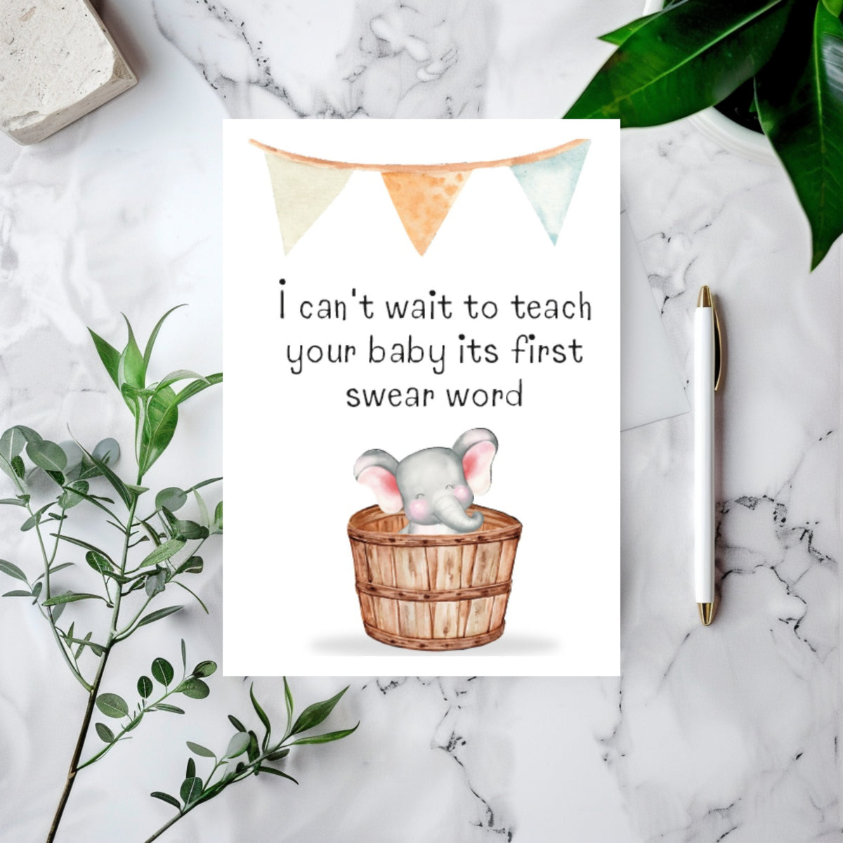 A baby shower greeting card featuring a watercolor elephant in a wooden basket with the text 'I can't wait to teach your baby its first swear word', styled in a flat lay on a white marble surface with green leaves, a pen, and a stone tile.