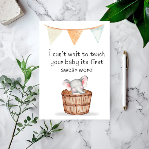 A baby shower greeting card featuring a watercolor elephant in a wooden basket with the text 'I can't wait to teach your baby its first swear word', styled in a flat lay on a white marble surface with green leaves, a pen, and a stone tile.