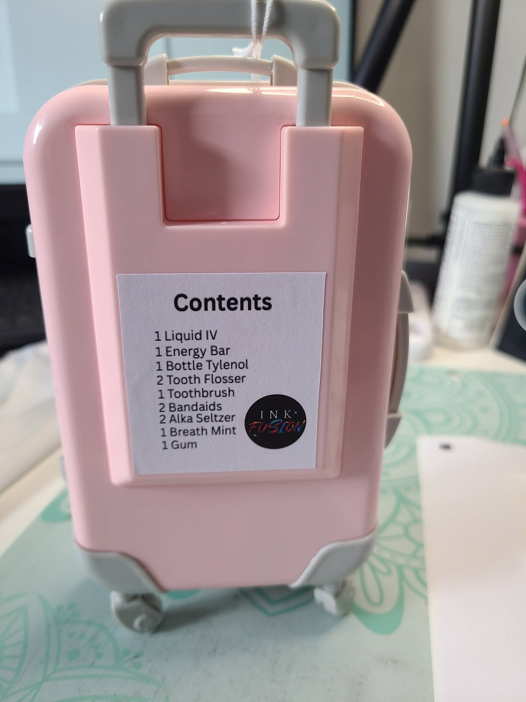 Back of a pink miniature suitcase with a contents label listing Liquid IV, an energy bar, Tylenol, tooth flossers, a toothbrush, bandages, Alka-Seltzer, a breath mint and gum.
