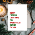 A white Christmas greeting card with bold red and green text reading 'Merry Fucking Christmas You Magnificent Mother Fucker', styled on a gray fabric surface with a white envelope, candle, pine branches, and holiday decor.